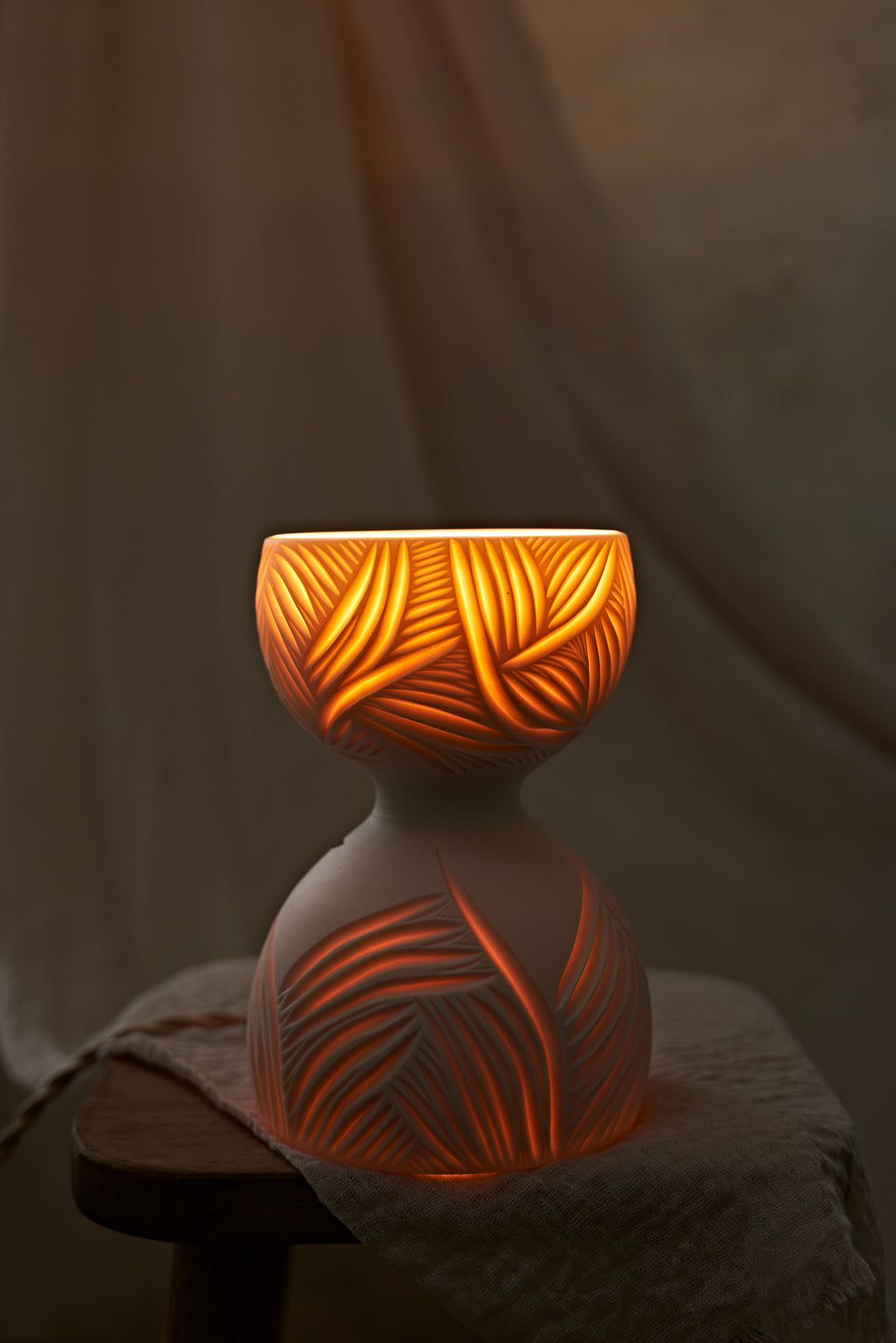 A white translucent porcelain ceramic hourglass-shaped table lamp with leaf-like carvings, glowing with warm orange light from within, displayed on a rustic wooden stool draped with a textured linen cloth against a soft beige fabric backdrop.