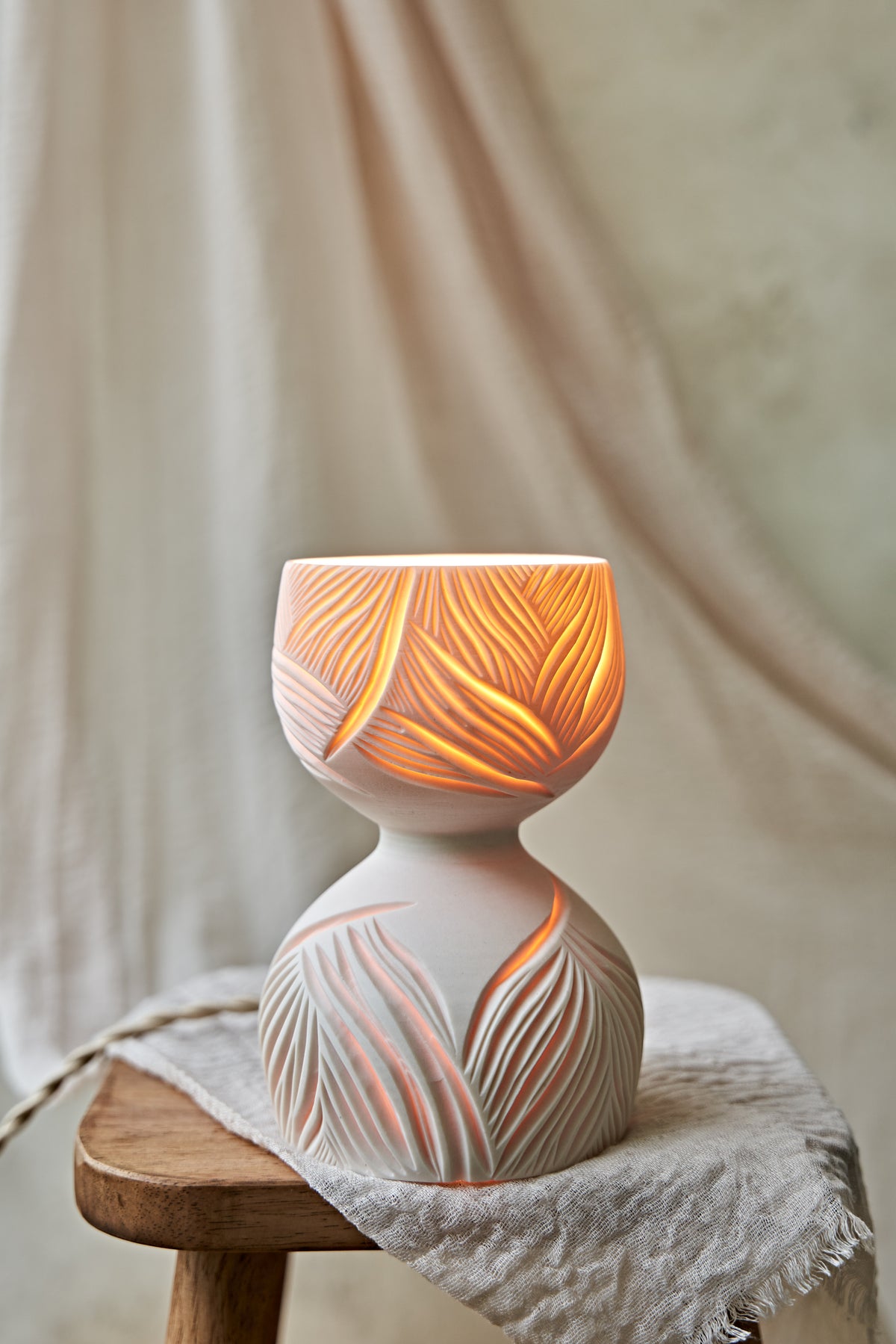 A porcelain ceramic table lamp with a matte off-white base and a glowing amber-orange top section featuring carved leaf-like patterns, displayed on a rustic wooden stool draped with linen.