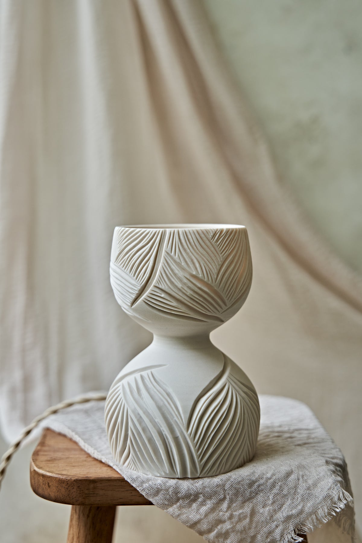 A porcelain ceramic table lamp with a matte off-white base and a glowing amber-orange top section featuring carved leaf-like patterns, displayed on a rustic wooden stool draped with linen.