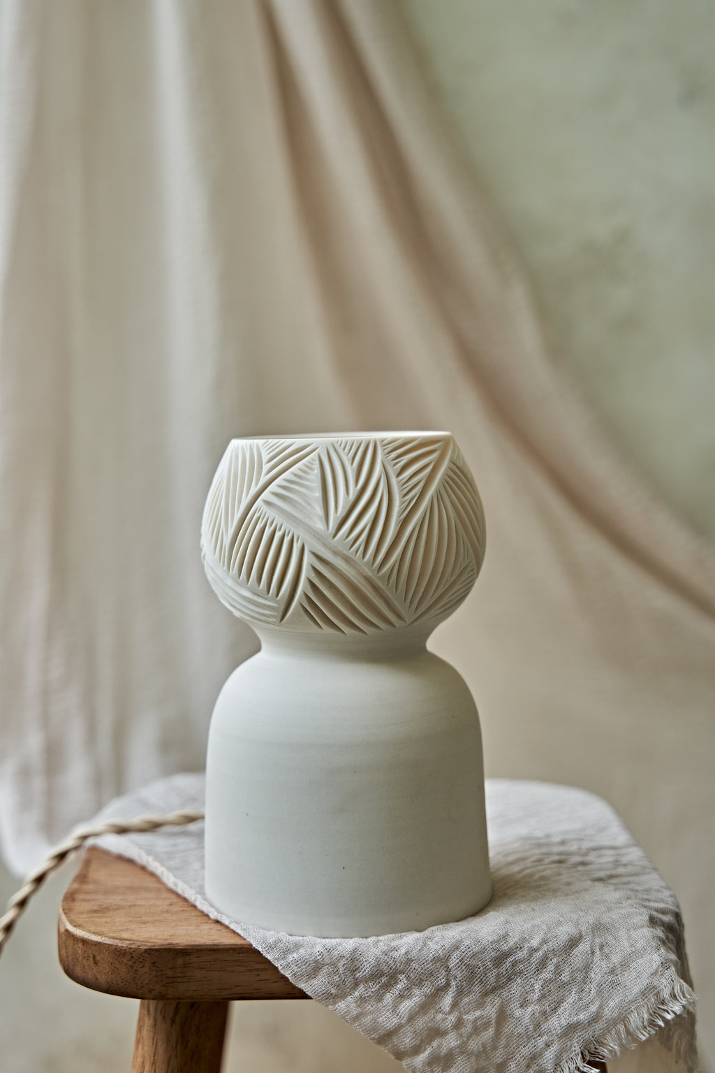 A porcelain ceramic table lamp with a matte off-white base and a glowing amber-orange top section when lit featuring carved leaf-like patterns, displayed on a rustic wooden stool draped with linen.