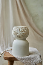 A porcelain ceramic table lamp with a matte off-white base and a glowing amber-orange top section when lit featuring carved leaf-like patterns, displayed on a rustic wooden stool draped with linen.