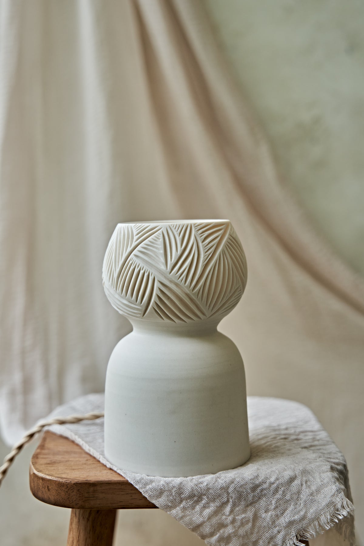 A porcelain ceramic table lamp with a matte off-white base and a glowing amber-orange top section when lit featuring carved leaf-like patterns, displayed on a rustic wooden stool draped with linen.