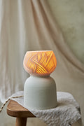A porcelain ceramic table lamp with a matte off-white base and a glowing amber-orange top section featuring carved leaf-like patterns, displayed on a rustic wooden stool draped with linen.