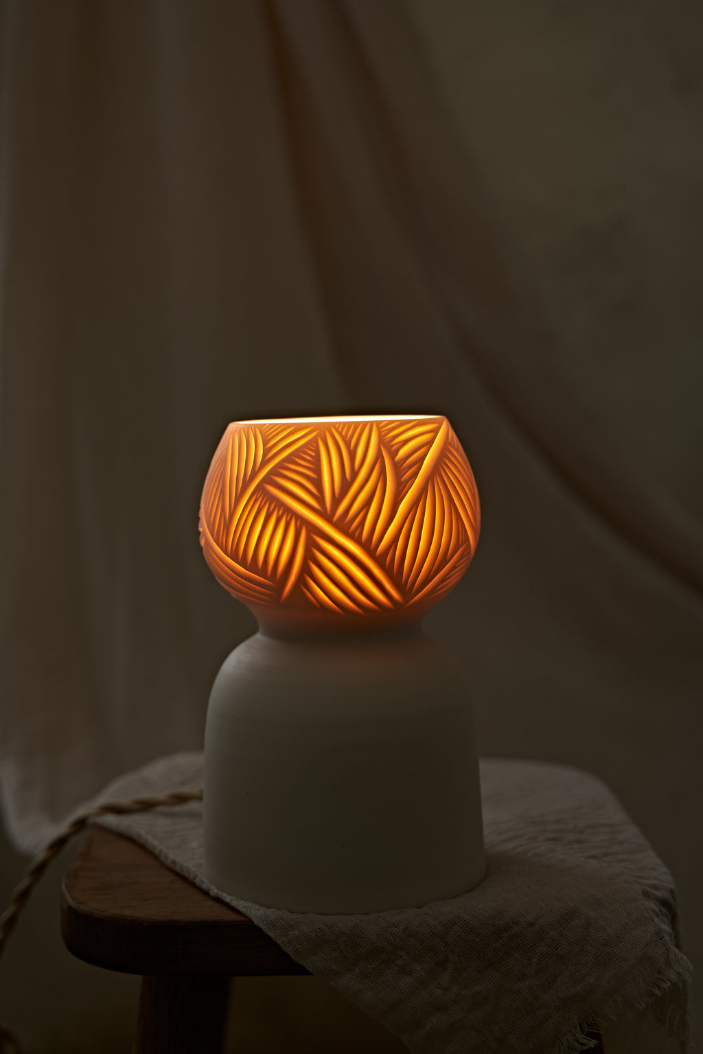 A porcelain ceramic table lamp with a matte off-white base and a glowing amber-orange top section featuring carved leaf-like patterns, displayed on a rustic wooden stool draped with linen.
