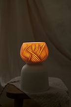 A porcelain ceramic table lamp with a matte off-white base and a glowing amber-orange top section featuring carved leaf-like patterns, displayed on a rustic wooden stool draped with linen.