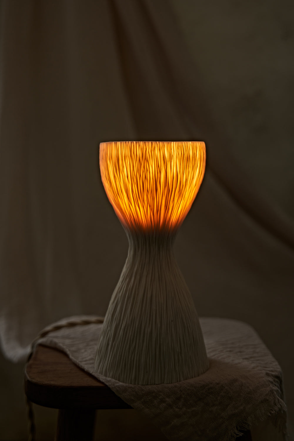 A textured, hourglass-shaped translucent porcelain table lamp with a ribbed white base and a glowing warm orange-yellow top, displayed on a rustic wooden stool draped with a light beige cloth.