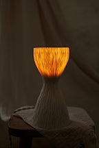 A textured, hourglass-shaped translucent porcelain table lamp with a ribbed white base and a glowing warm orange-yellow top, displayed on a rustic wooden stool draped with a light beige cloth.