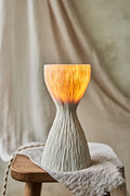 A textured, hourglass-shaped translucent porcelain table lamp with a ribbed white base and a glowing warm orange-yellow top, displayed on a rustic wooden stool draped with a light beige cloth.