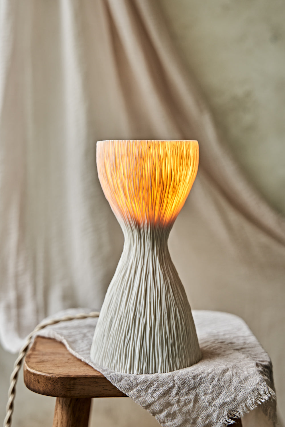 A textured, hourglass-shaped translucent porcelain table lamp with a ribbed white base and a glowing warm orange-yellow top, displayed on a rustic wooden stool draped with a light beige cloth.