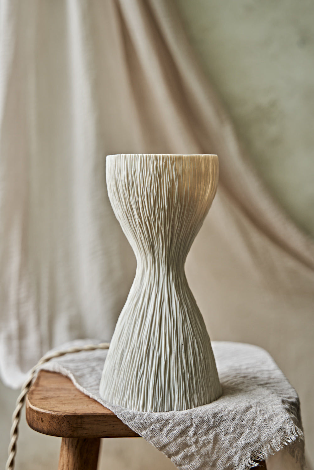 A textured, hourglass-shaped translucent porcelain table lamp with a ribbed white base and a glowing warm orange-yellow top, displayed on a rustic wooden stool draped with a light beige cloth.