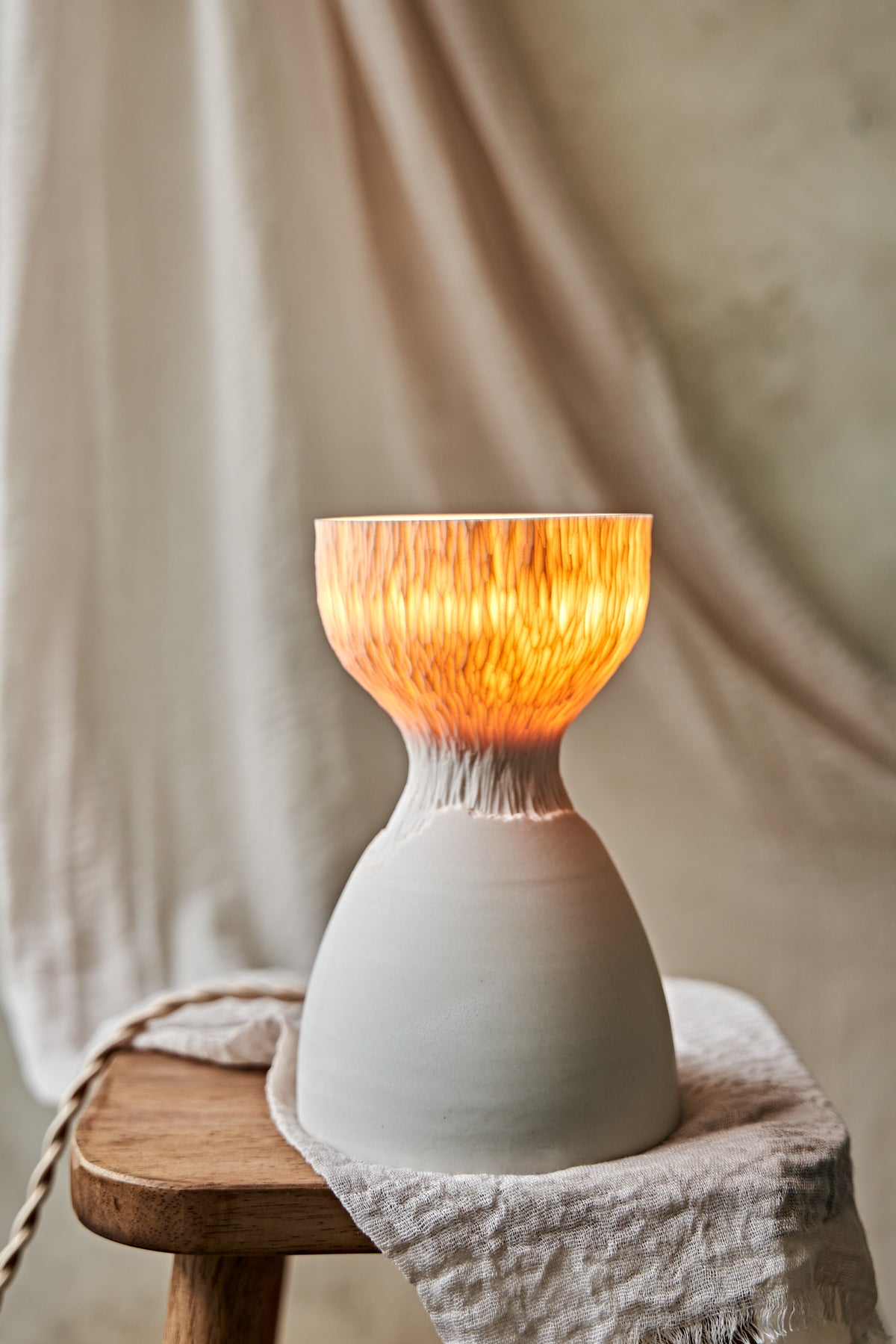 A modern ceramic table lamp with a matte white base and a flared carved amber-orange glowing top when lit, styled on a rustic wooden stool with draped linen fabric.

