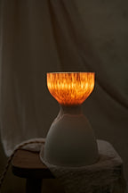 A modern ceramic table lamp with a matte white base and a flared carved amber-orange glowing top when lit, styled on a rustic wooden stool with draped linen fabric.