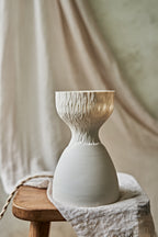 A modern ceramic table lamp with a matte white base and a flared carved amber-orange glowing top when lit, styled on a rustic wooden stool with draped linen fabric.