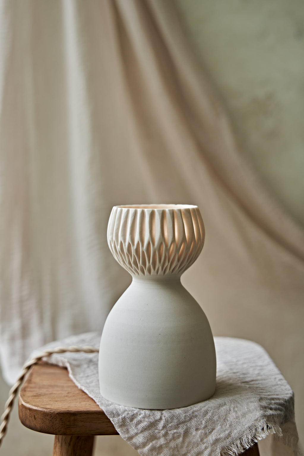 A translucent porcelain ceramic table lamp with a matte off-white body and a ribbed, fluted glass shade glowing warm orange, displayed on a rustic wooden stool with draped linen in a softly blurred indoor setting.