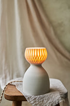 A translucent porcelain ceramic table lamp with a matte off-white body and a ribbed, fluted glass shade glowing warm orange, displayed on a rustic wooden stool with draped linen in a softly blurred indoor setting.
