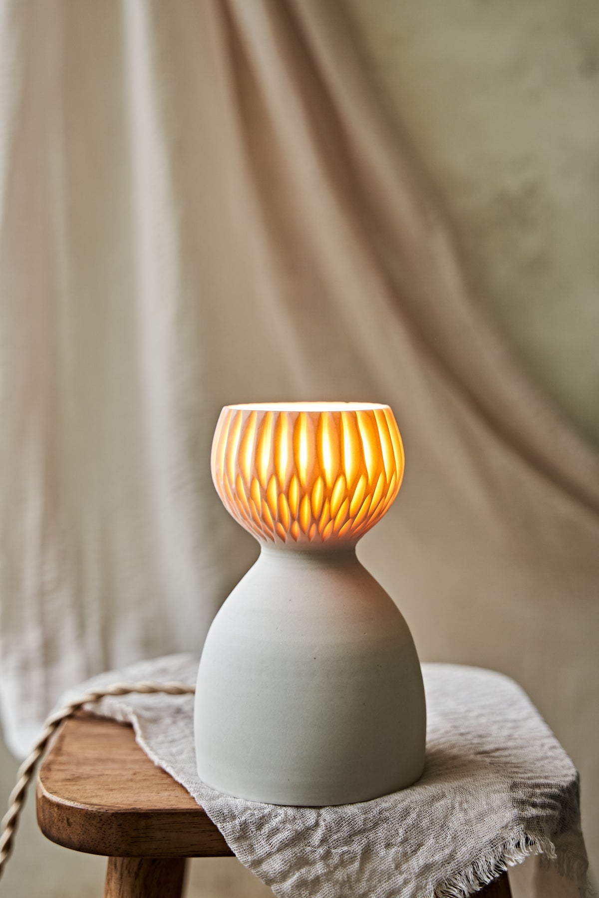 A translucent porcelain ceramic table lamp with a matte off-white body and a ribbed, fluted glass shade glowing warm orange, displayed on a rustic wooden stool with draped linen in a softly blurred indoor setting.