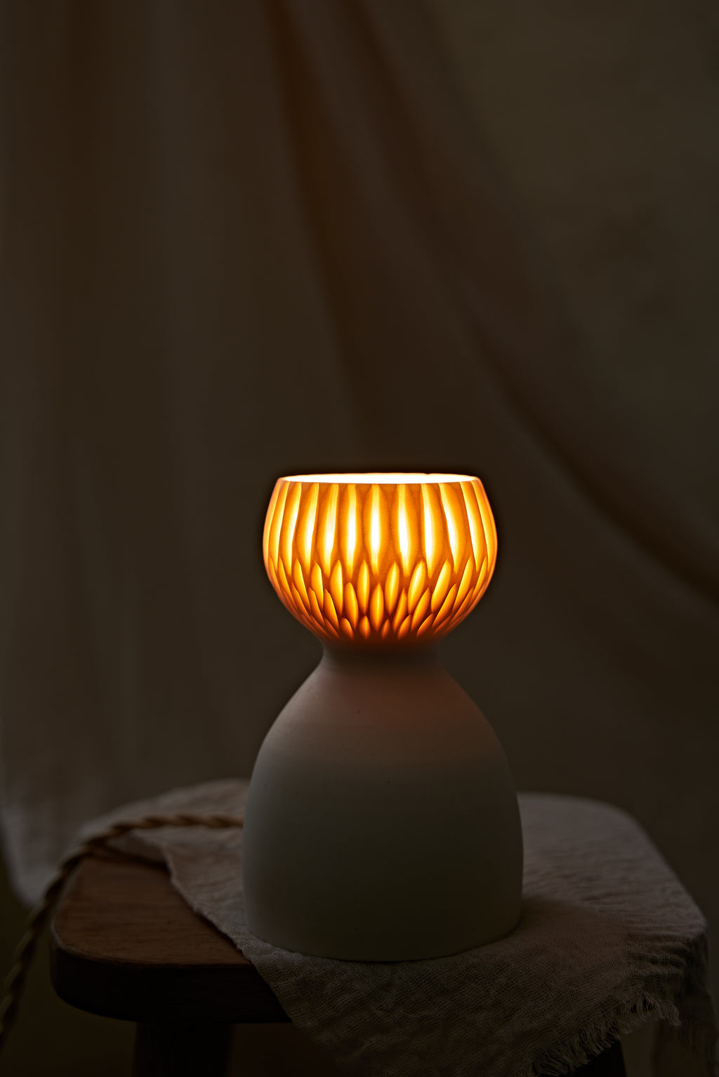 A translucent porcelain ceramic table lamp with a matte off-white body and a ribbed, fluted glass shade glowing warm orange, displayed on a rustic wooden stool with draped linen in a softly blurred indoor setting.