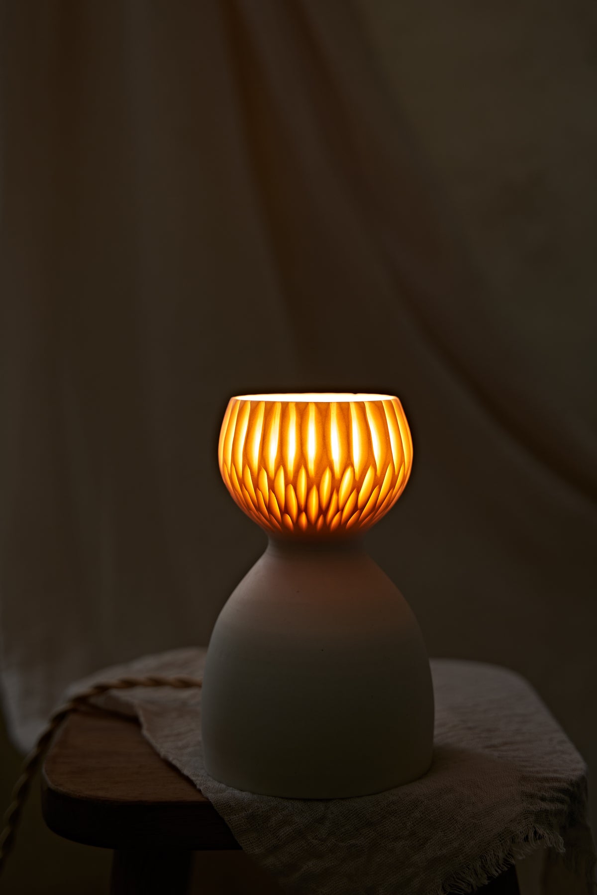 A translucent porcelain ceramic table lamp with a matte off-white body and a ribbed, fluted glass shade glowing warm orange, displayed on a rustic wooden stool with draped linen in a softly blurred indoor setting.