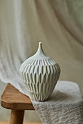 A ribbed ceramic vase in a light cream color, displayed on a rustic wooden stool draped with a neutral linen cloth against a soft beige background.