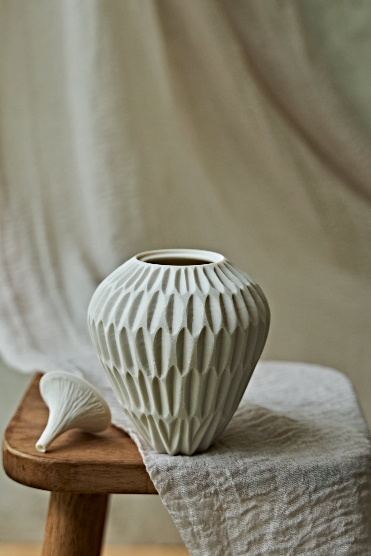 A ribbed ceramic vase in a warm off-white or cream color, styled on a rustic wooden stool draped with a linen cloth.