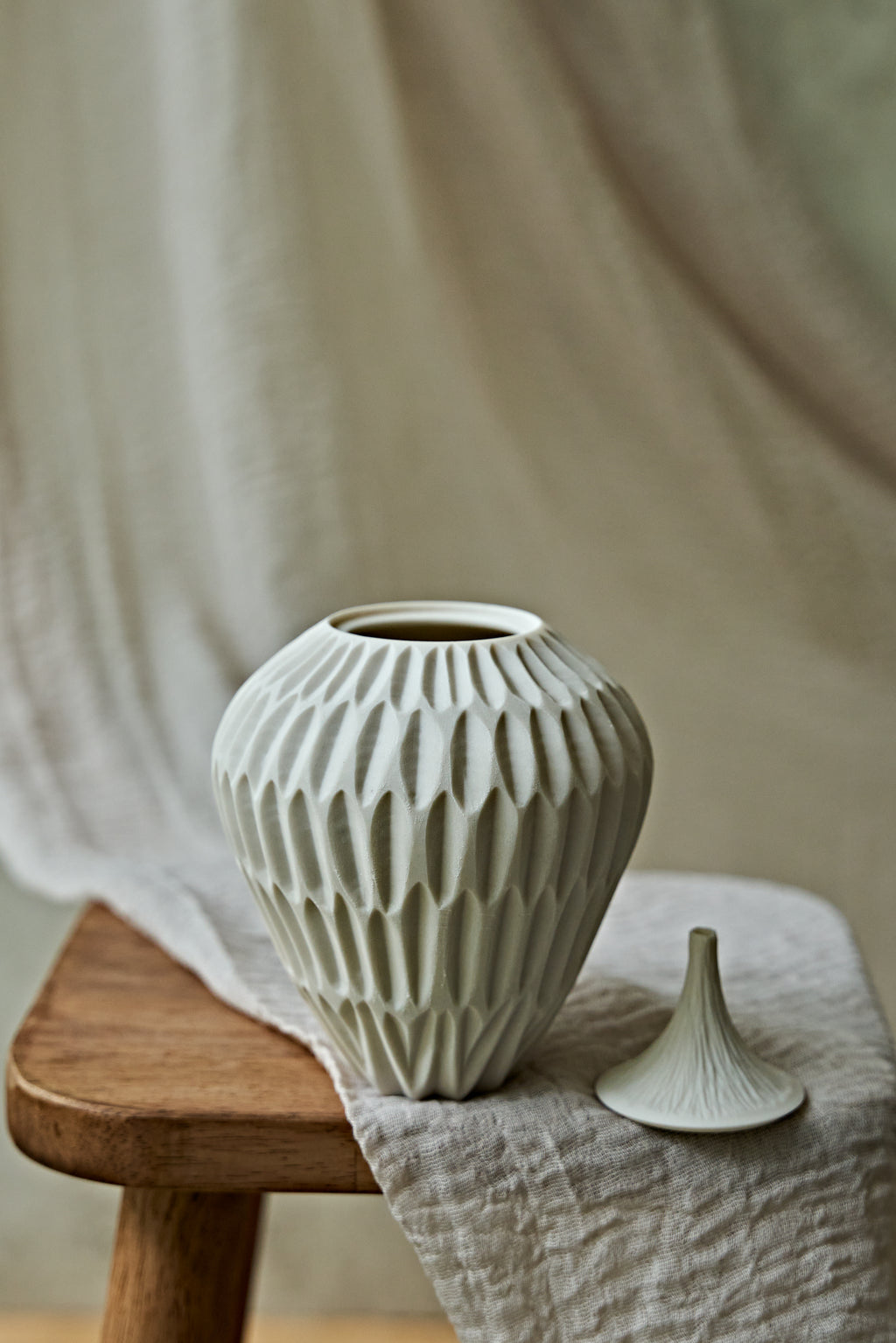 A ribbed ceramic vase in a light warm gray or off-white glaze sits on a wooden stool beside its matching lid, draped in neutral fabric against a softly blurred background.