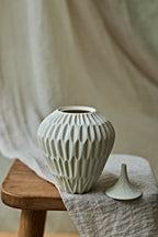 A ribbed ceramic vase in a light warm gray or off-white glaze sits on a wooden stool beside its matching lid, draped in neutral fabric against a softly blurred background.