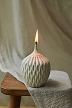A textured ceramic oil lamp with a lit flame, resting on a wooden stool draped with a neutral linen cloth.