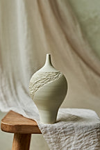 A light cream porcelain ceramic oil lamp with a textured, ribbed pattern on the upper half, displayed on a rustic wooden stool draped with a neutral linen cloth.