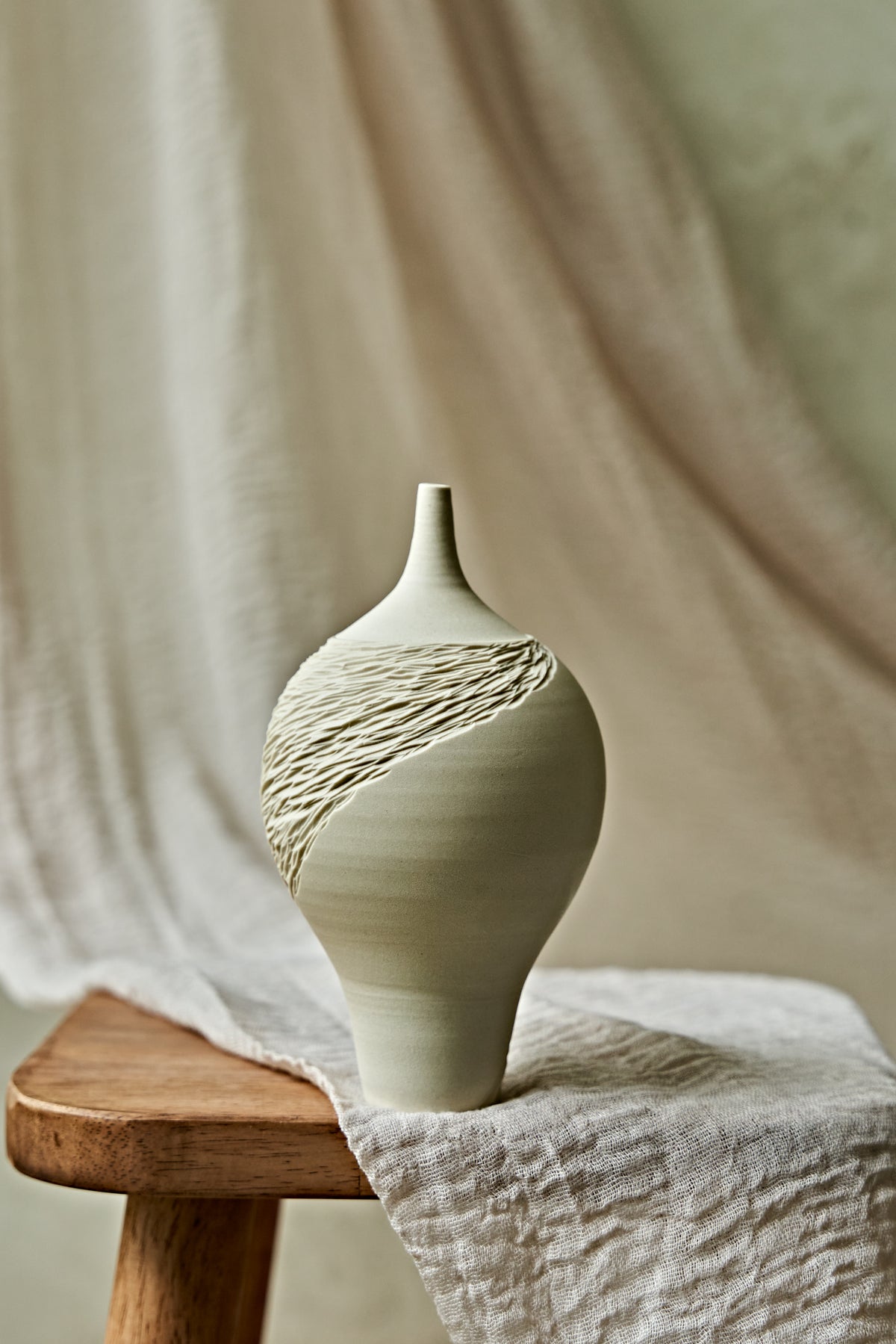 A light cream porcelain ceramic oil lamp with a textured, ribbed pattern on the upper half, displayed on a rustic wooden stool draped with a neutral linen cloth.