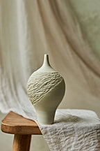 A light cream porcelain ceramic oil lamp with a textured, ribbed pattern on the upper half, displayed on a rustic wooden stool draped with a neutral linen cloth.
