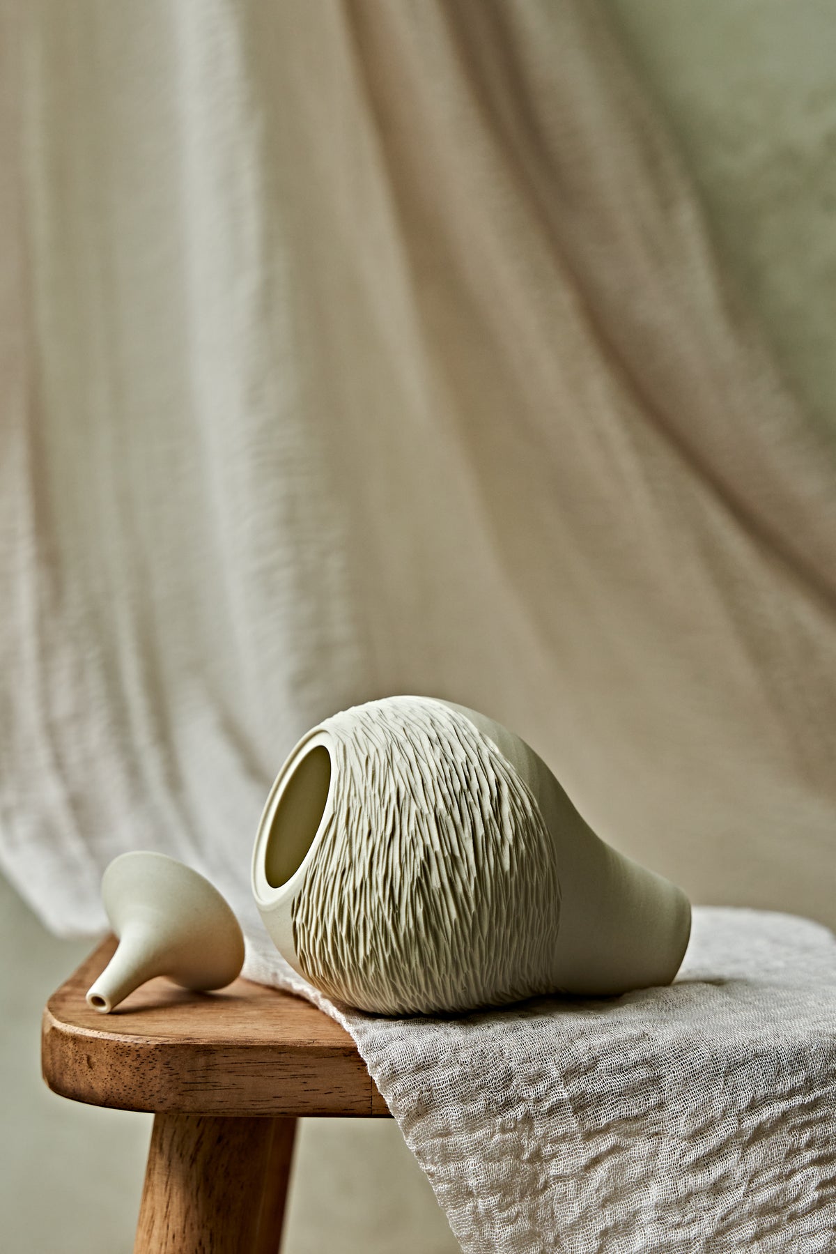 A light cream porcelain ceramic oil lamp with a textured, ribbed pattern on the upper half, displayed on a rustic wooden stool draped with a neutral linen cloth.