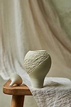 A light cream porcelain ceramic oil lamp with a textured, ribbed pattern on the upper half, displayed on a rustic wooden stool draped with a neutral linen cloth.