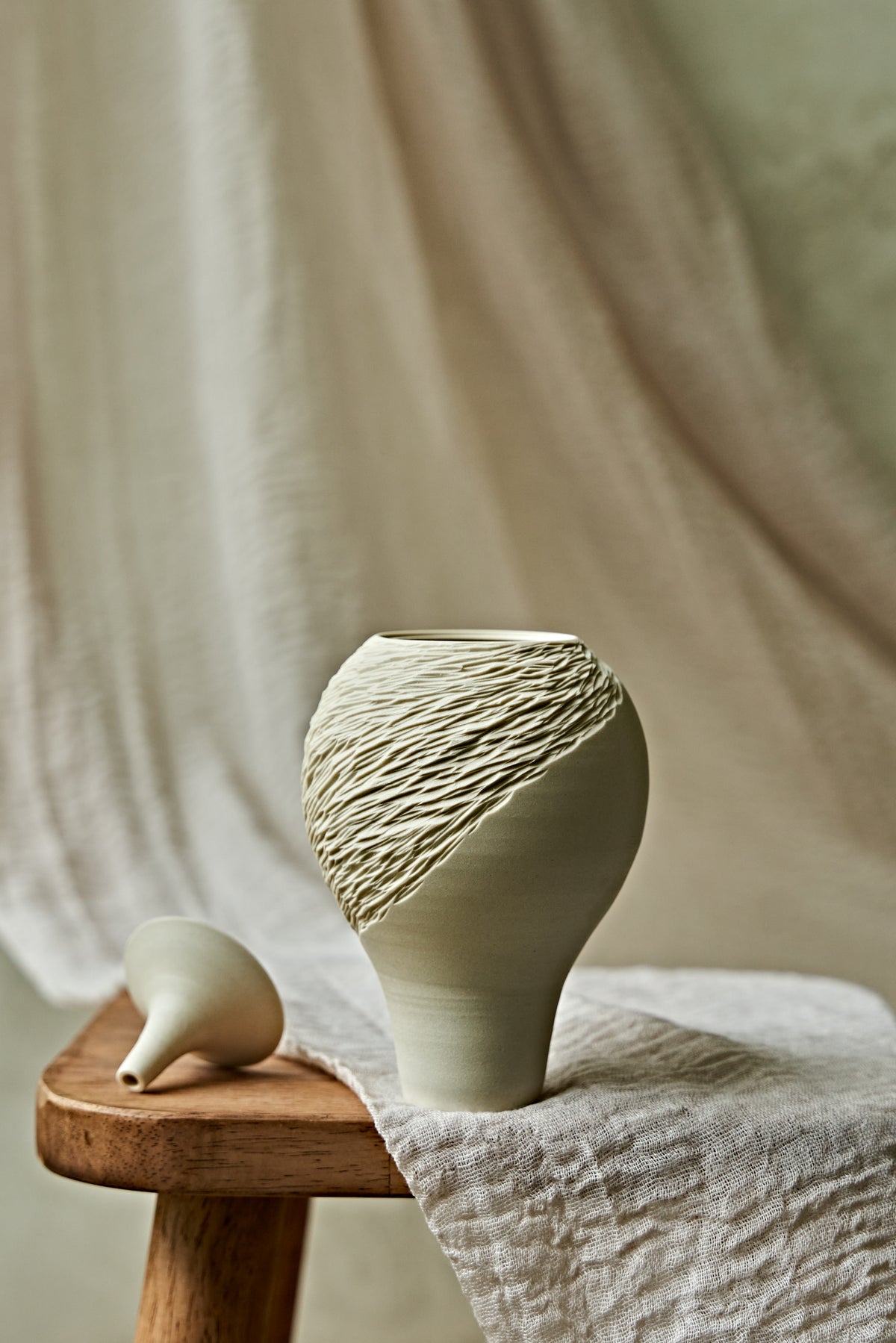 A light cream porcelain ceramic oil lamp with a textured, ribbed pattern on the upper half, displayed on a rustic wooden stool draped with a neutral linen cloth.