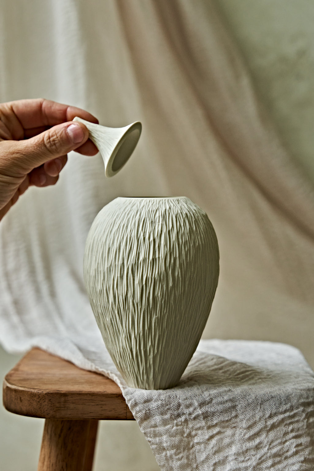 A textured ceramic porcelain oil lamp in a light cream color, displayed on a rustic wooden stool draped with a neutral linen cloth against a soft, draped fabric backdrop.