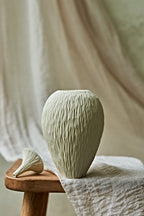 A textured ceramic porcelain oil lamp in a light cream color, displayed on a rustic wooden stool draped with a neutral linen cloth against a soft, draped fabric backdrop.