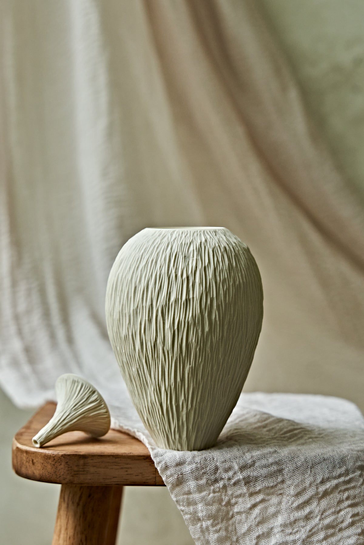 A textured ceramic porcelain oil lamp in a light cream color, displayed on a rustic wooden stool draped with a neutral linen cloth against a soft, draped fabric backdrop.