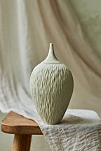 A textured ceramic porcelain oil lamp in a light cream color, displayed on a rustic wooden stool draped with a neutral linen cloth against a soft, draped fabric backdrop.