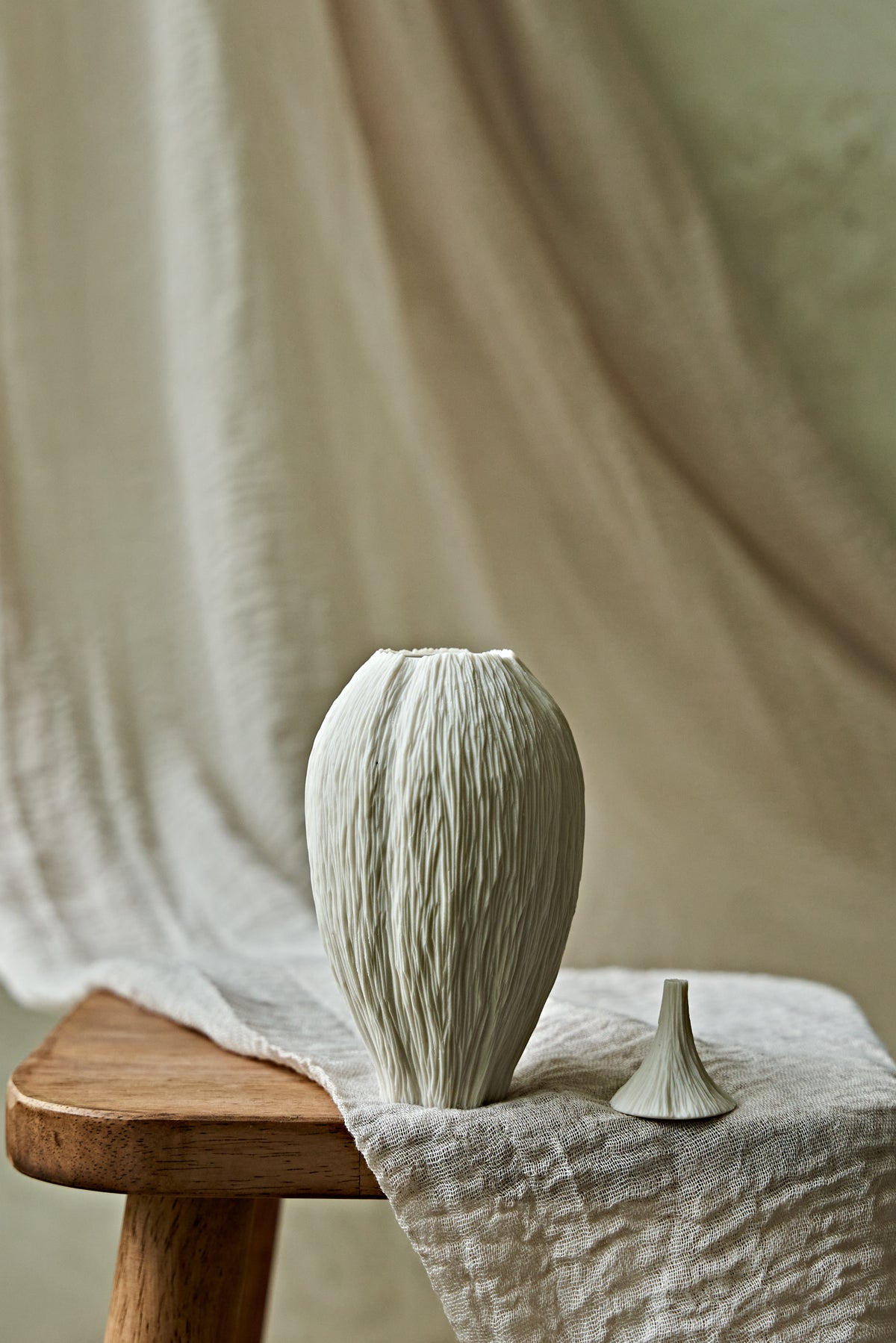 A textured ceramic porcelain oil lamp in a warm off-white tone stands on a wooden stool draped with linen fabric, alongside a small matching decorative object, against a soft beige draped backdrop.