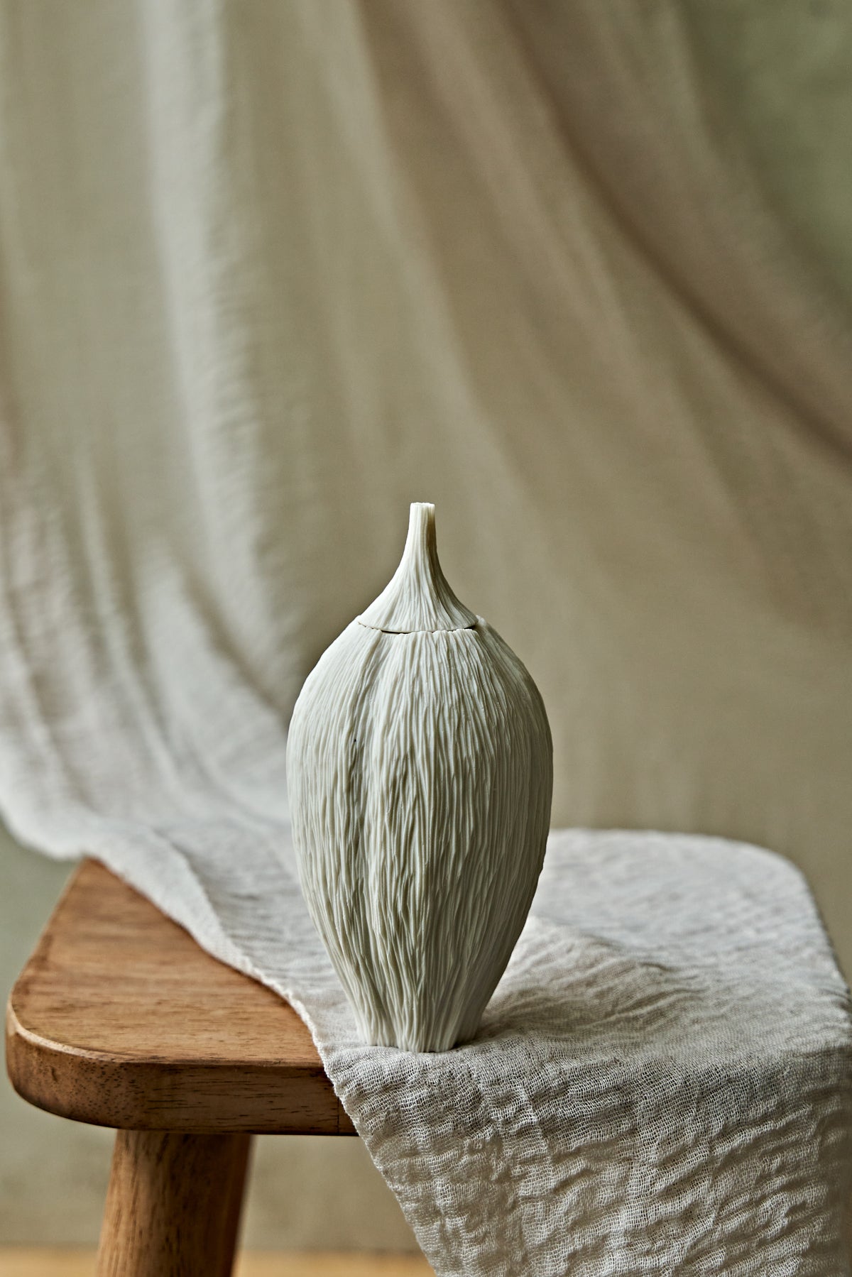 A textured ceramic porcelain oil lamp in a warm off-white tone stands on a wooden stool draped with linen fabric, alongside a small matching decorative object, against a soft beige draped backdrop.