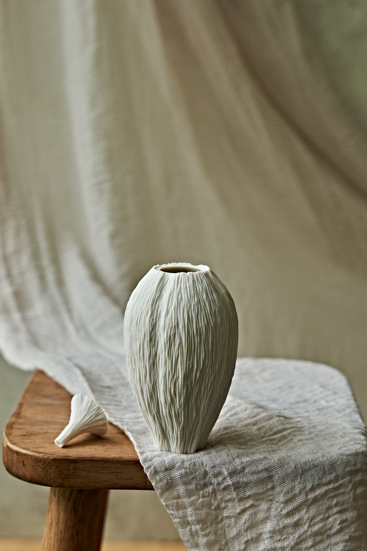 A textured ceramic porcelain oil lamp in a warm off-white tone stands on a wooden stool draped with linen fabric, alongside a small matching decorative object, against a soft beige draped backdrop.