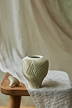 A small ceramic porcelain oil lamp with a narrow neck and carved leaf-like texture, resting on a wooden stool draped with light fabric against a soft neutral backdrop.