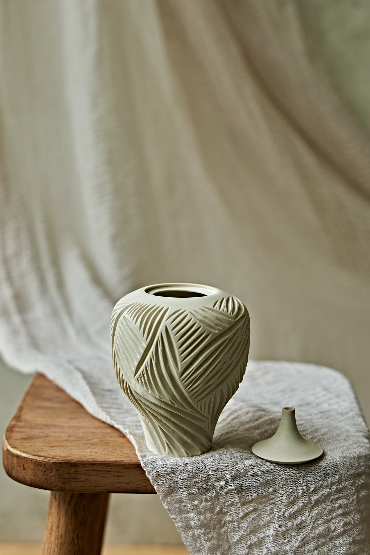 A small ceramic porcelain oil lamp with a narrow neck and carved leaf-like texture, resting on a wooden stool draped with light fabric against a soft neutral backdrop.