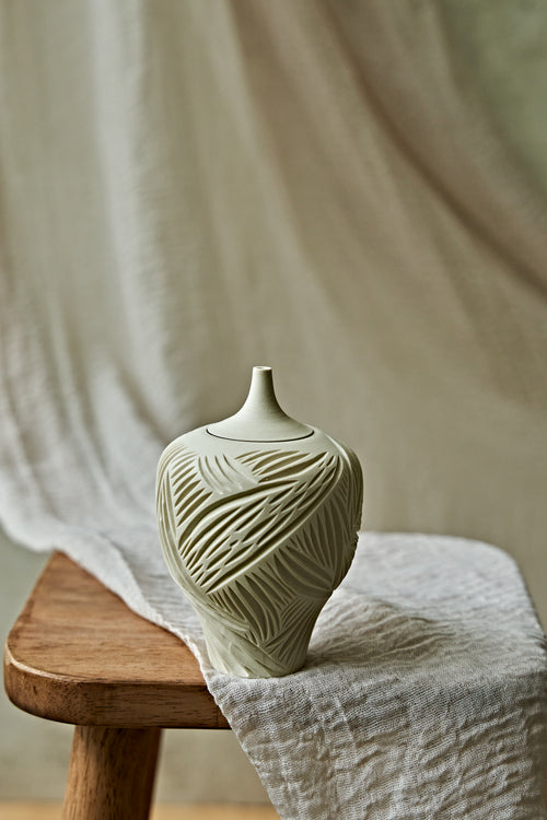 A small ceramic porcelain oil lamp with a narrow neck and carved leaf-like texture, resting on a wooden stool draped with light fabric against a soft neutral backdrop.

