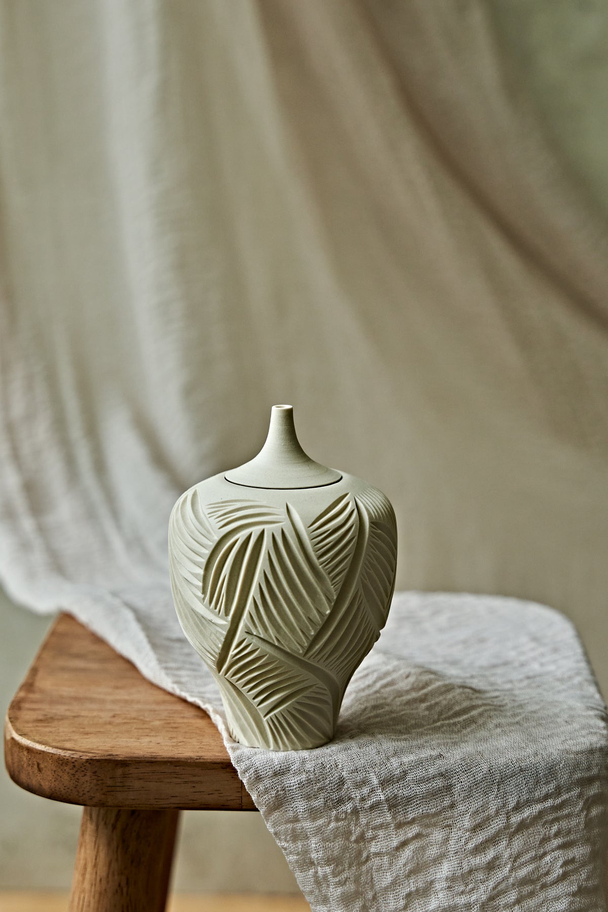 A small ceramic porcelain oil lamp with a narrow neck and carved leaf-like texture, resting on a wooden stool draped with light fabric against a soft neutral backdrop.