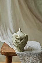 A small ceramic porcelain oil lamp with a narrow neck and carved leaf-like texture, resting on a wooden stool draped with light fabric against a soft neutral backdrop.