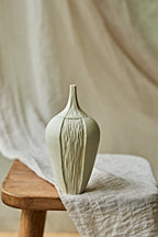 A light cream ceramic oil lamp with a textured vertical ribbed pattern, displayed on a rustic wooden stool draped with a neutral linen cloth.
