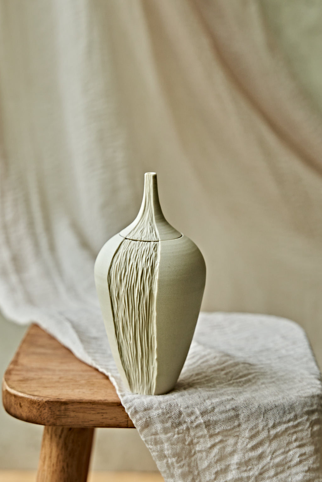 A light cream ceramic oil lamp with a textured vertical ribbed pattern, displayed on a rustic wooden stool draped with a neutral linen cloth.