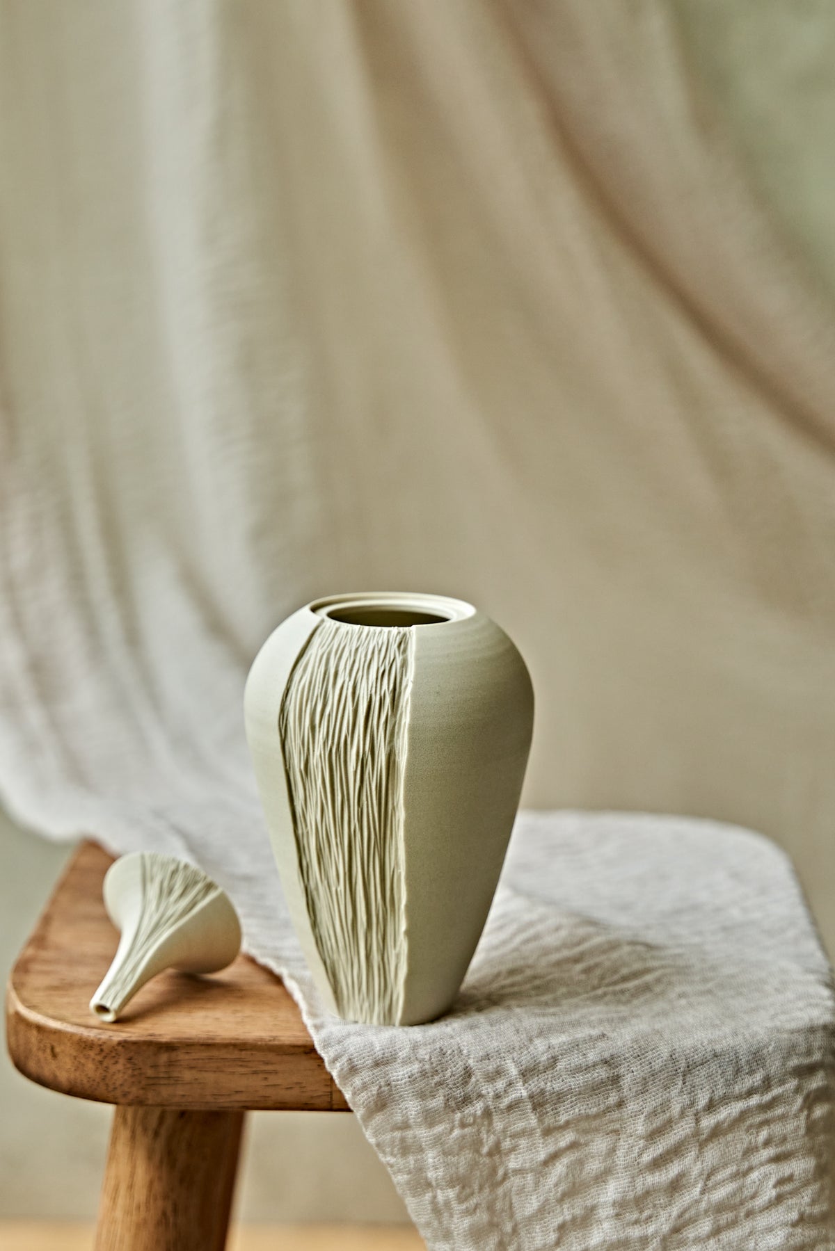 A light cream ceramic oil lamp with a textured vertical ribbed pattern, displayed on a rustic wooden stool draped with a neutral linen cloth.