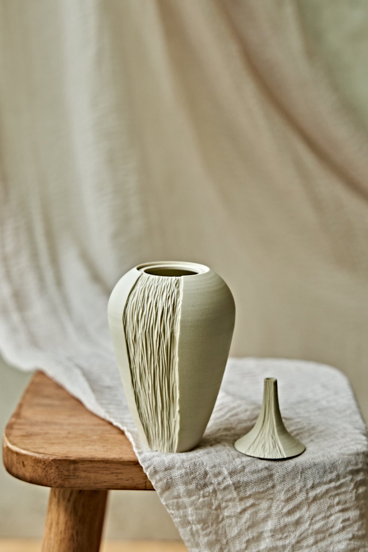 A light cream ceramic oil lamp with a textured vertical ribbed pattern, displayed on a rustic wooden stool draped with a neutral linen cloth.