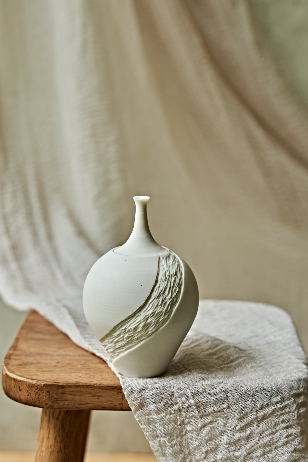 A small matte white ceramic porcelain oil lamp with a textured diagonal wave design, displayed on a rustic wooden stool draped with a neutral linen cloth.

